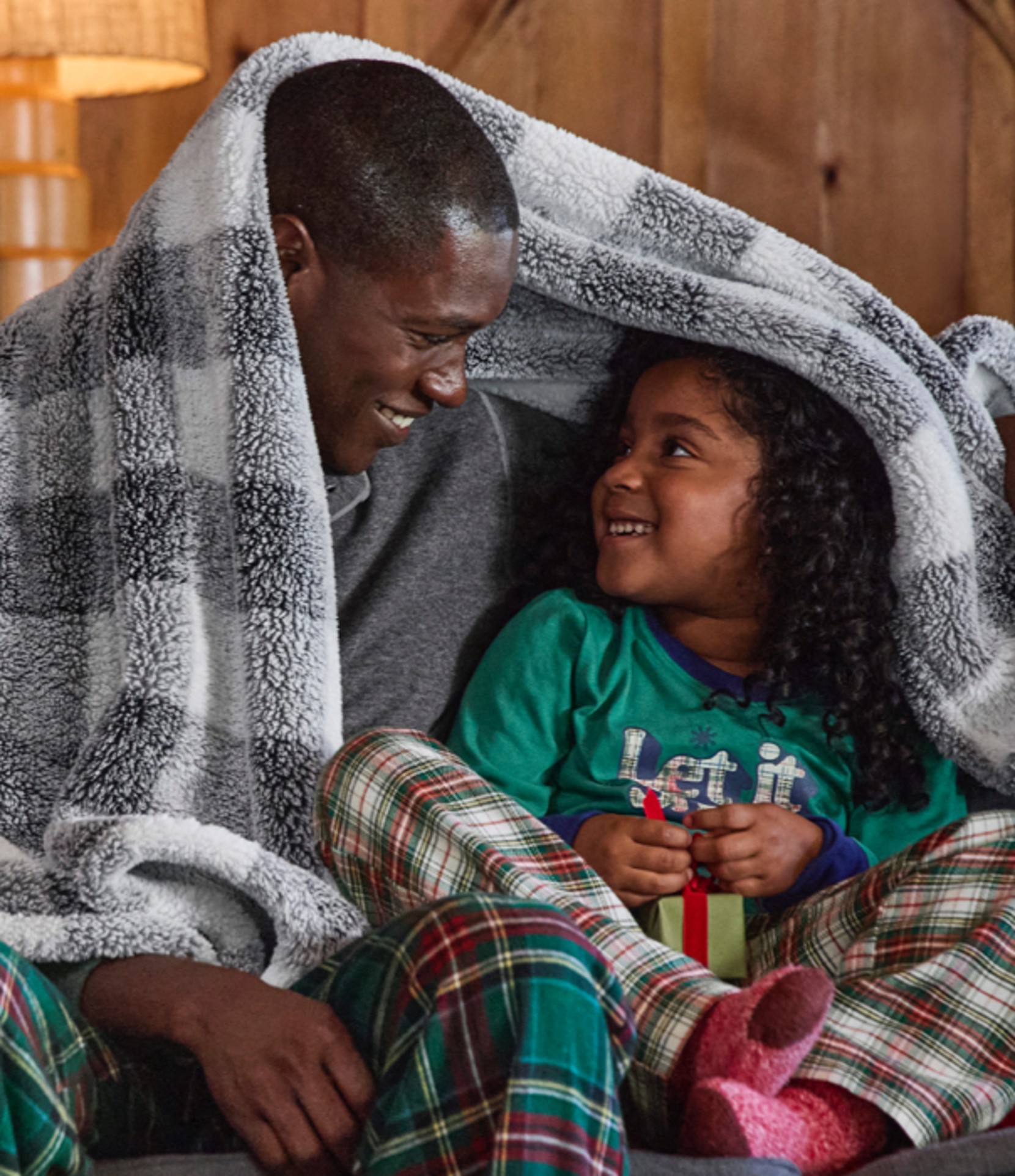 Two people in plaid pajamas sit on a couch, sharing a checkered blanket, with a lamp and wooden wall behind them.