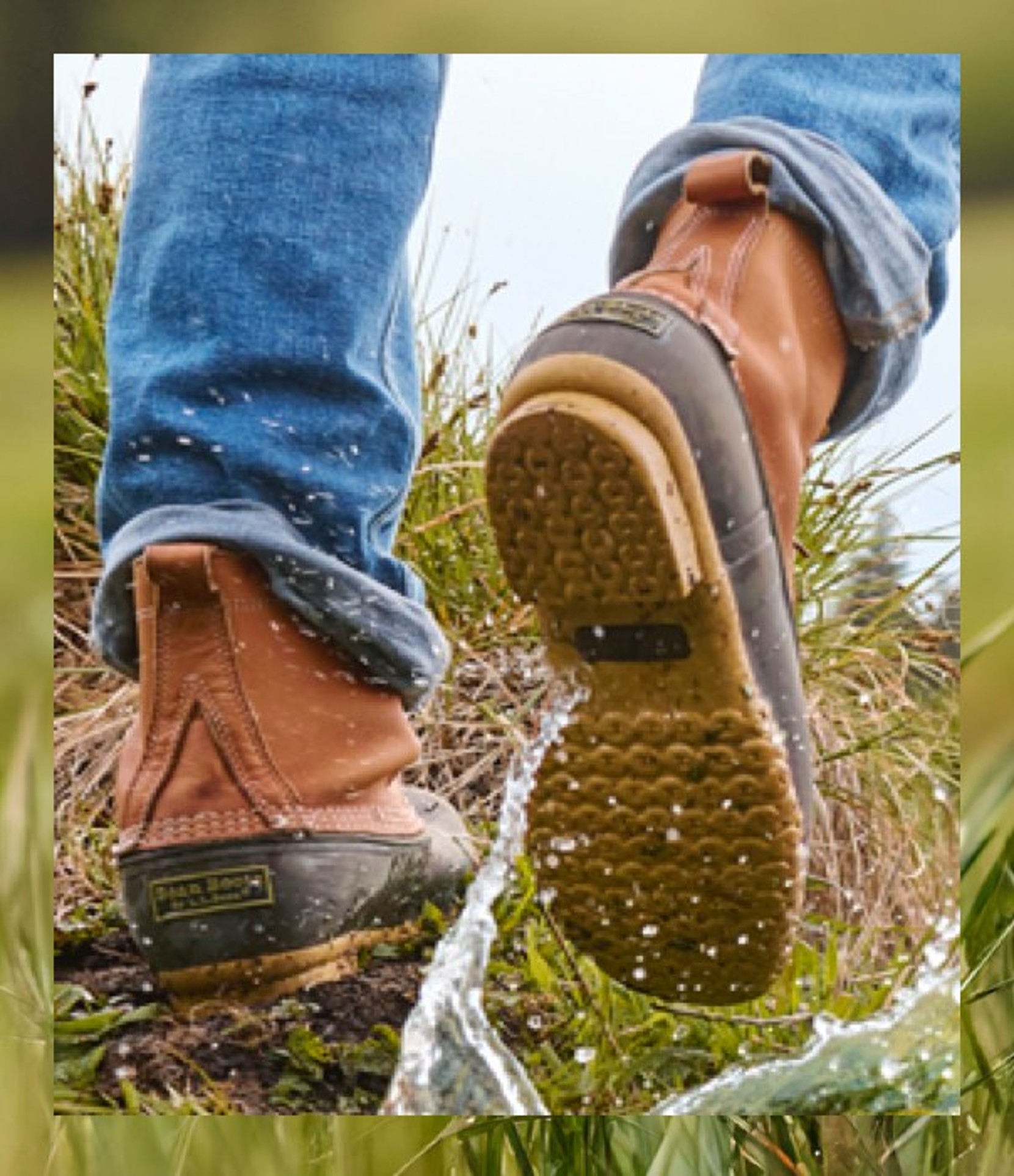 Close-up of boots splashing through water on grassy terrain, person wearing rolled-up jeans.