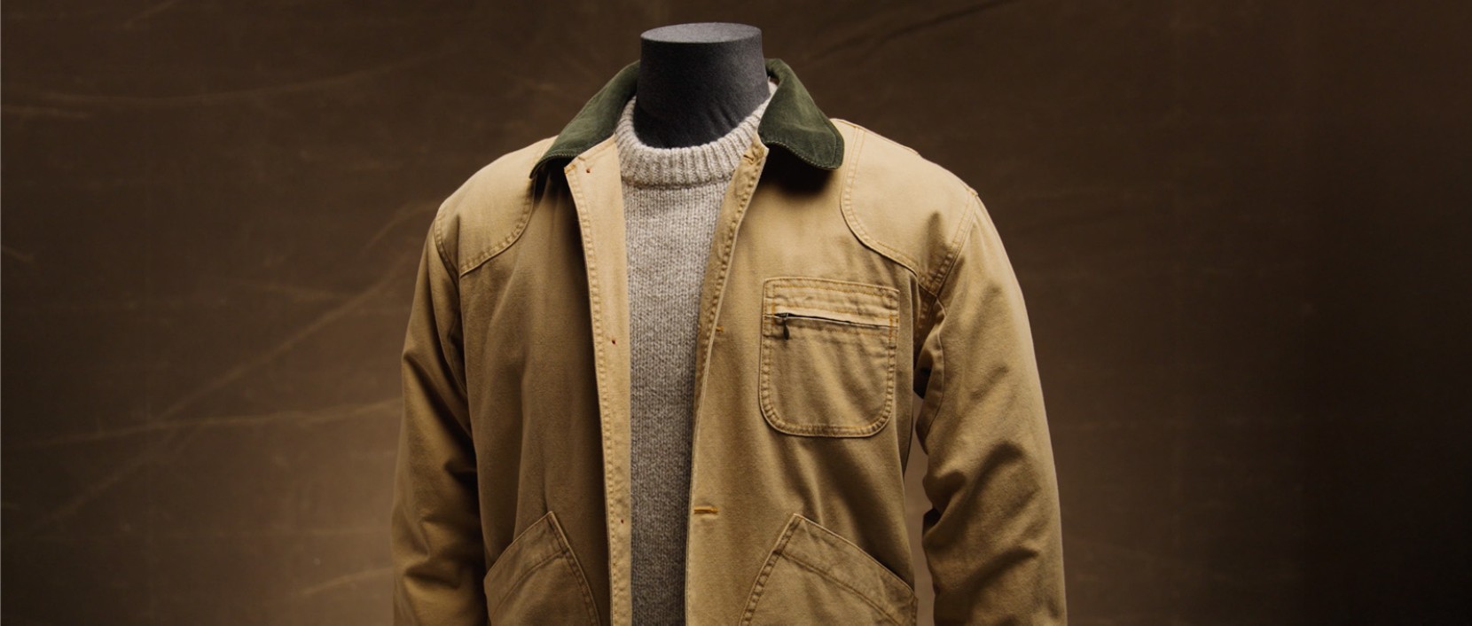 Tan field coat with a green collar shown on a mannequin over a gray sweater against a brown backdrop.