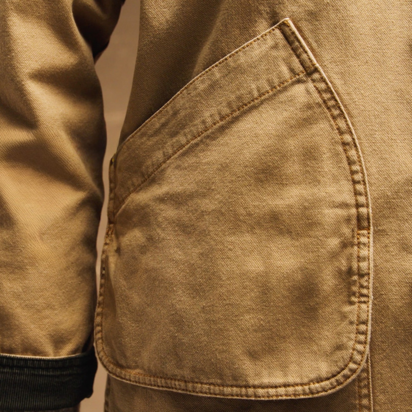 Close up of a tan field coat’s large front pocket and sleeve, showing stitching and worn fabric texture.