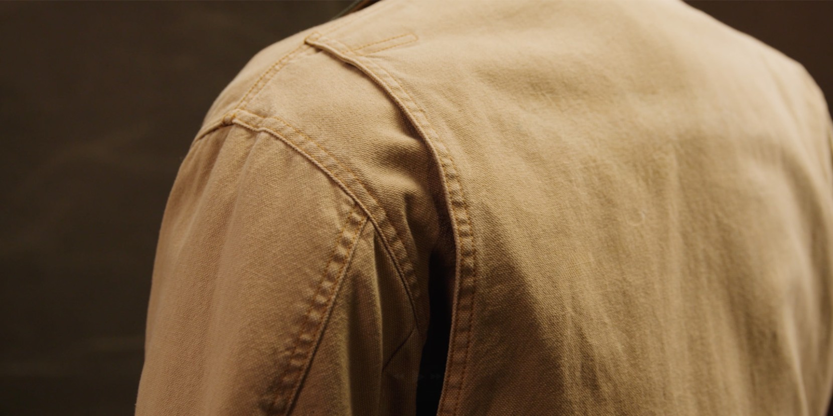 Tan field coat with a green collar shown on a mannequin over a gray sweater against a brown backdrop.