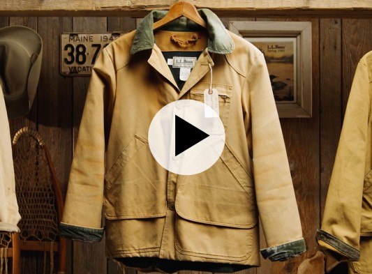 Vintage tan L L Bean coat on a hanger in a rustic room with gear and décor in the background.