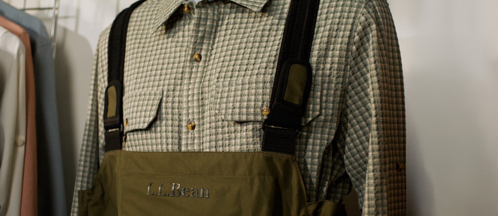 Close-up of olive L.L.Bean chest waders with black suspenders worn over a green check button-down shirt on a rack.