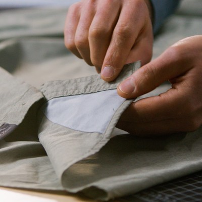 Hands pulling open a shirt pocket to show a light-colored interior lining sewn into the garment.