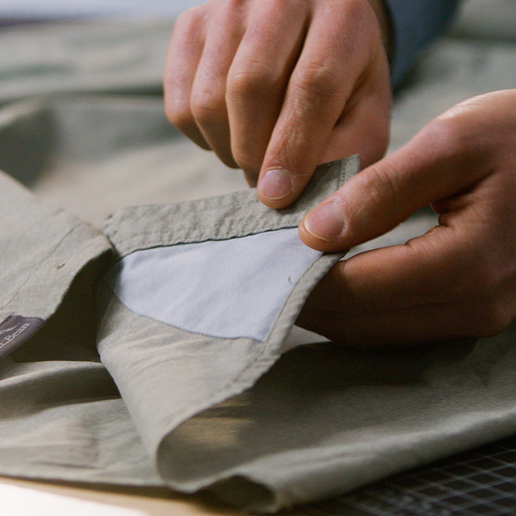 Hands pulling open a shirt pocket to show a light-colored interior lining sewn into the garment.