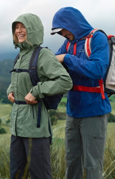 Two people in hooded rain jackets adjust their backpacks while standing in a grassy outdoor setting.