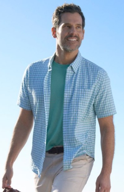 Person wearing a teal T‑shirt with a light blue short‑sleeve checkered shirt and light pants, walking outdoors under a clear sky.