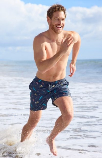Shirtless man running through shallow ocean water, wearing dark patterned swim shorts under a bright sky.