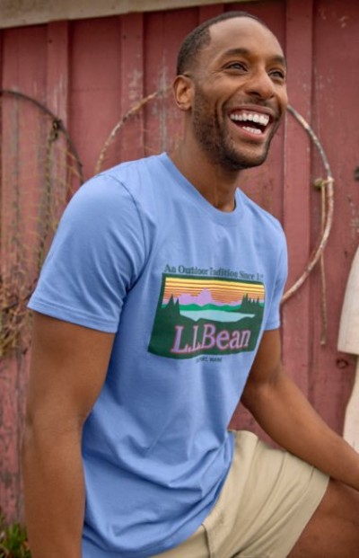 Person wearing a light blue L.L.Bean graphic T‑shirt and khaki shorts while sitting in front of a weathered red wall.
