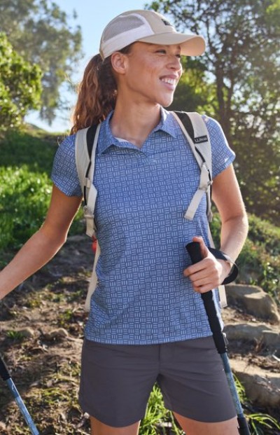 Wearing a cap, backpack, and hiking gear, the person holds trekking poles on a trail surrounded by greenery.