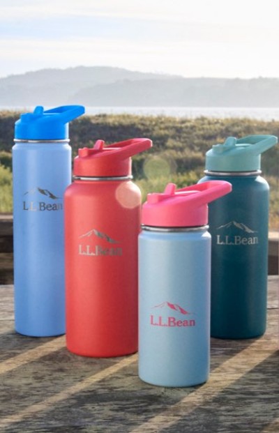 Four colorful L. L. Bean water bottles with flip-top lids arranged on a wooden surface outdoors near a scenic shoreline.