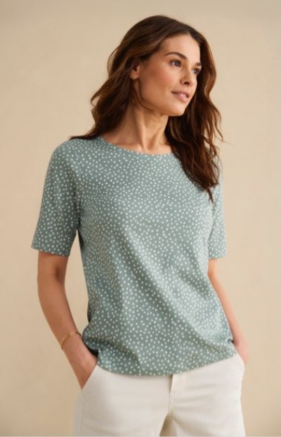 Wearing a patterned short‑sleeve top and light pants, a person stands with hands in pockets against a neutral background.