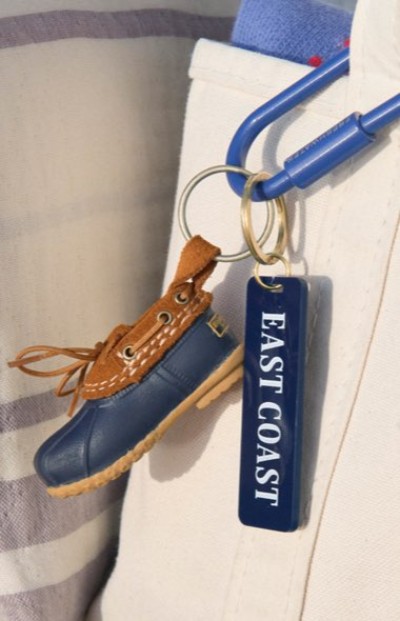 Mini boot‑shaped keychain and an “EAST COAST” tag hanging from a blue carabiner clipped to light-colored canvas bag.