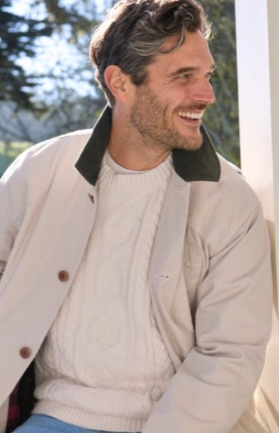 Person wearing a light jacket and cable‑knit sweater sits outdoors near a porch with trees in the background.