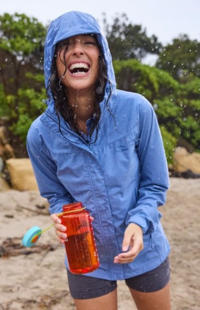 Wearing a blue hooded jacket and shorts, a person stands laughing in the rain holding a large orange water bottle.