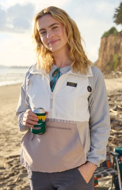 Standing on a beach at sunset, a person holds a canned drink and wears a light pullover with a coastal backdrop.