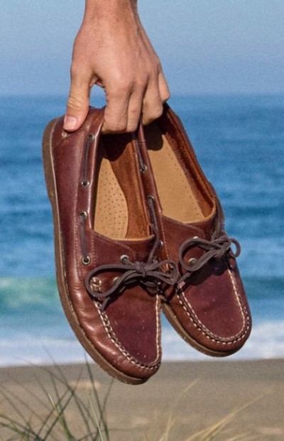 Close up of a hand holding a pair of brown leather moccasins with laces against a beach and ocean backdrop.
