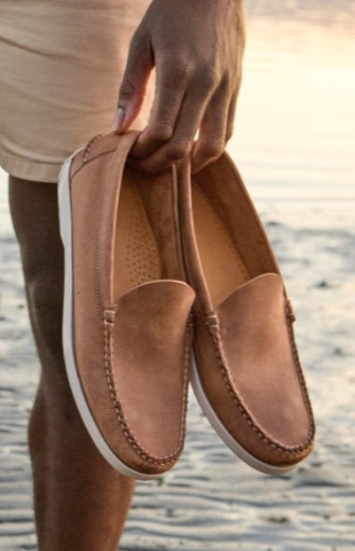 Close up image of a hand holding a pair of brown slip‑on shoes while standing on a sandy beach.
