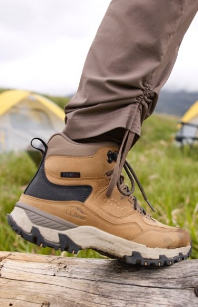 Person in brown hiking boots and pants steps on a log in a grassy outdoor area with tents visible in the background.