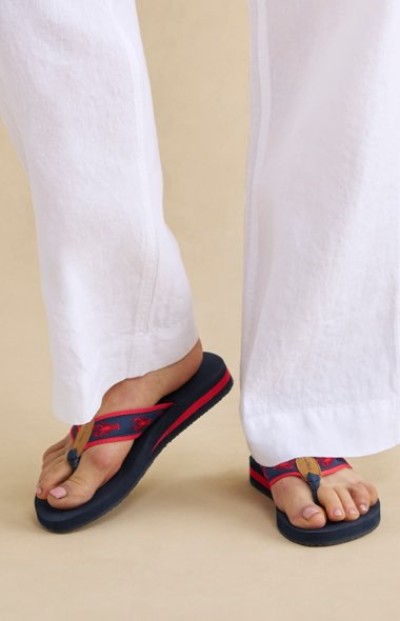 Navy flip‑flops with red straps worn with loose white pants, shown against a simple beige background.