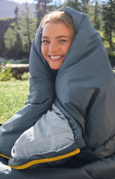 A person wrapped in a large gray sleeping bag sits outdoors on a grassy area with trees and mountains in the background.