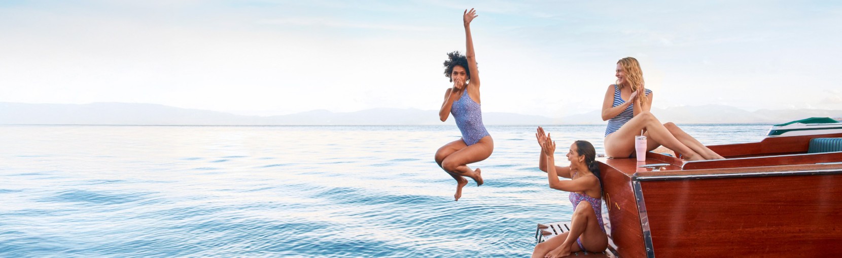 Friends on a wooden boat laugh and clap as one jumps into a calm lake, with distant mountains under a clear sky.