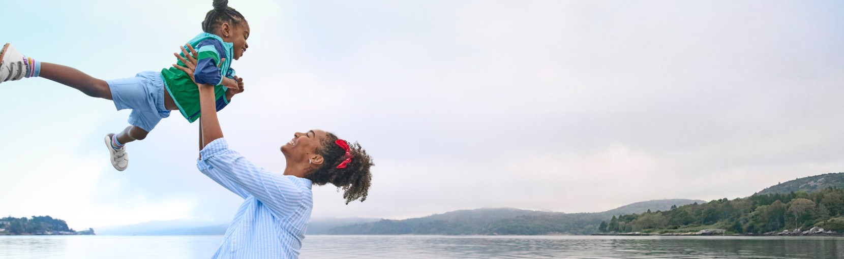 An adult lifts a child into the air near a calm lakeshore, with hills and water stretching across the background.