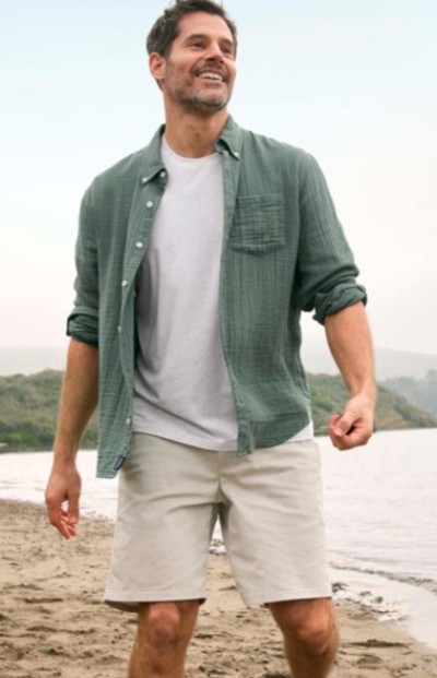 Smiling man wearing a white T‑shirt layered under an open, green button-up shirt walking outdoors near water.