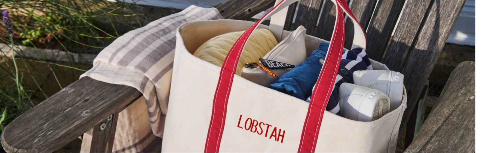 Canvas tote with red straps labeled “LOBSTAH,” filled with shoes, towel, clothing, and sunscreen on a wooden chair outdoors.