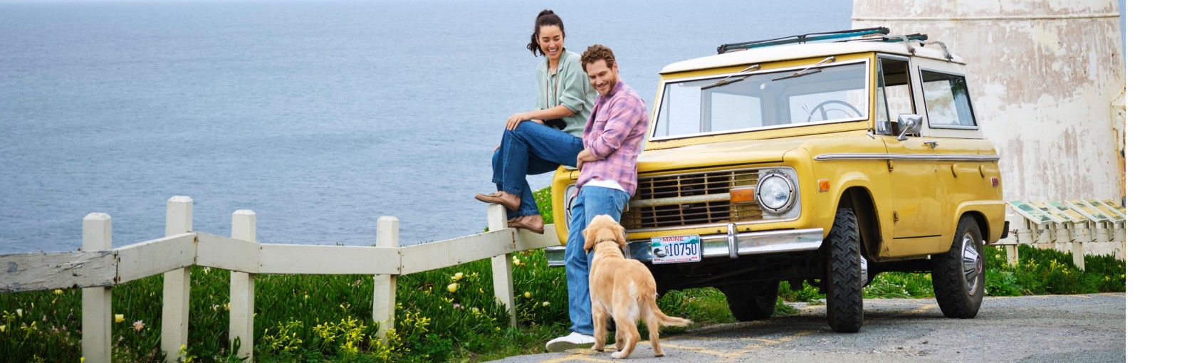 Two people sit by a coastal fence beside a yellow vintage SUV, with a dog standing nearby and ocean cliffs in the background.