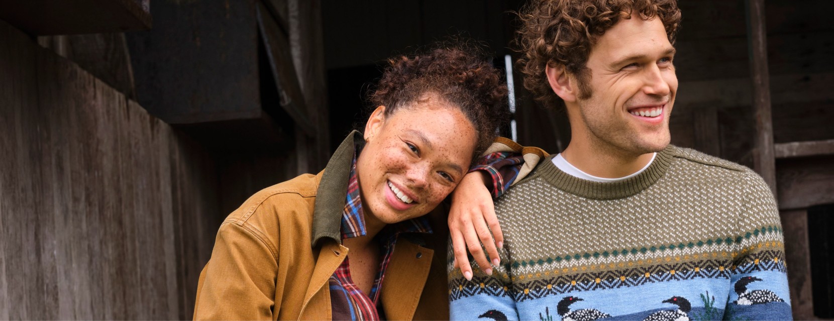 Two people in fall layers pose outdoors by a rustic wooden building, one wearing a patterned loon sweater.