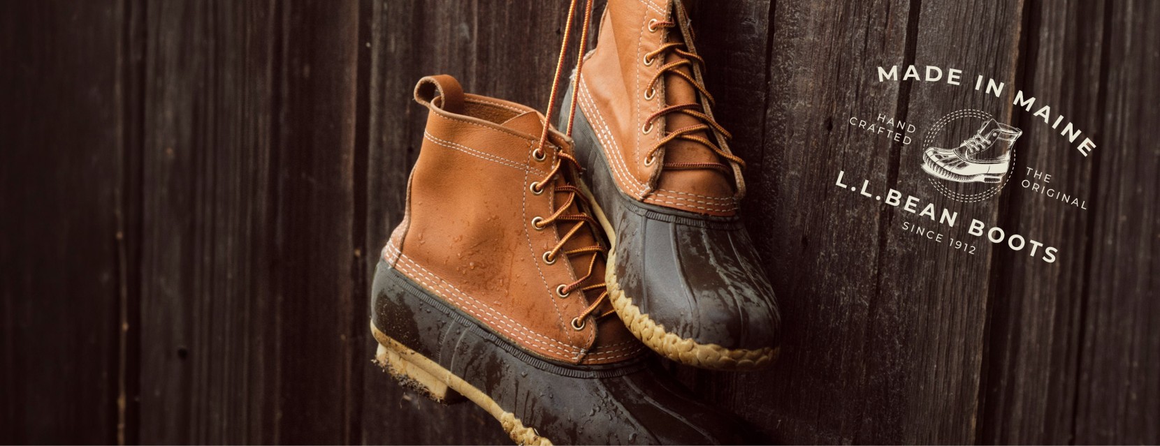 Pair of L. L. Bean boots hang by their laces against a dark wooden wall with text Made in Maine since 1912.