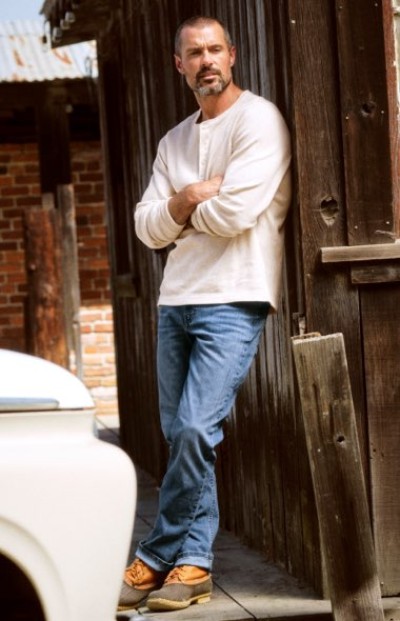 Person standing in a rustic doorway wearing a cream shirt, jeans, and work boots.