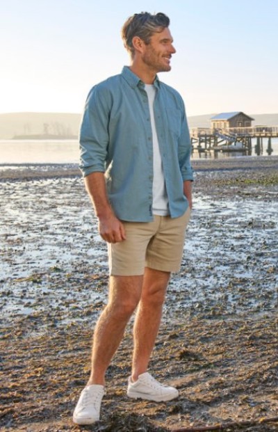 Person standing on a beach wearing a blue button-down shirt, shorts, and sneakers.