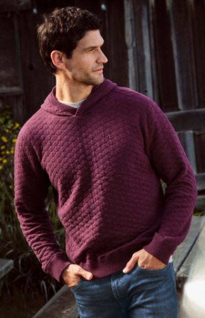Person wearing a burgundy knit sweater and jeans standing outside near a rustic building.