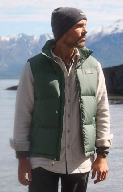Person wearing layers under a green insulated vest and knit hat stands beside a lake and mountains.
