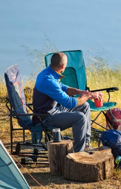 Camper seated beside a tent and campfire setup in a grassy field with water in the distance.