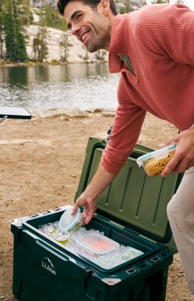 Person organizing food containers inside a green cooler at a lakeside beach.