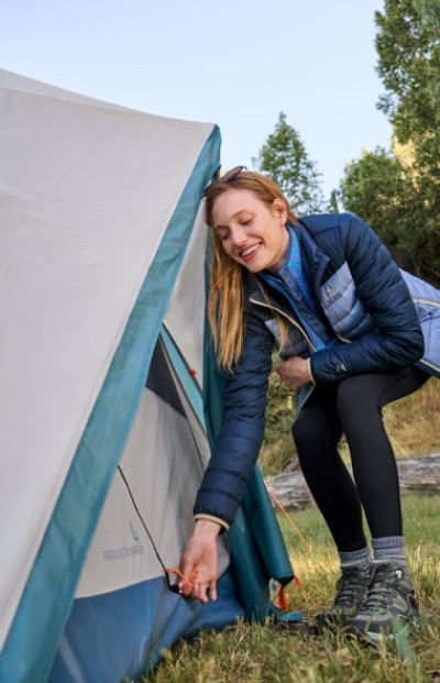 Person zipping up a blue and gray tent at a campsite with greenery behind.