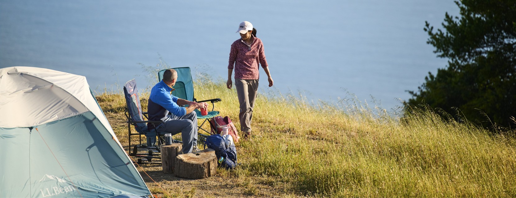 Family campsite with a tent, chairs and campers in an open field near water.