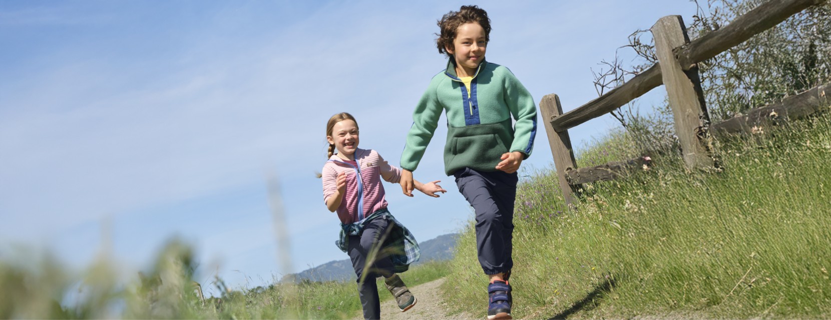 Two children run through a grassy field on a sunny day.
