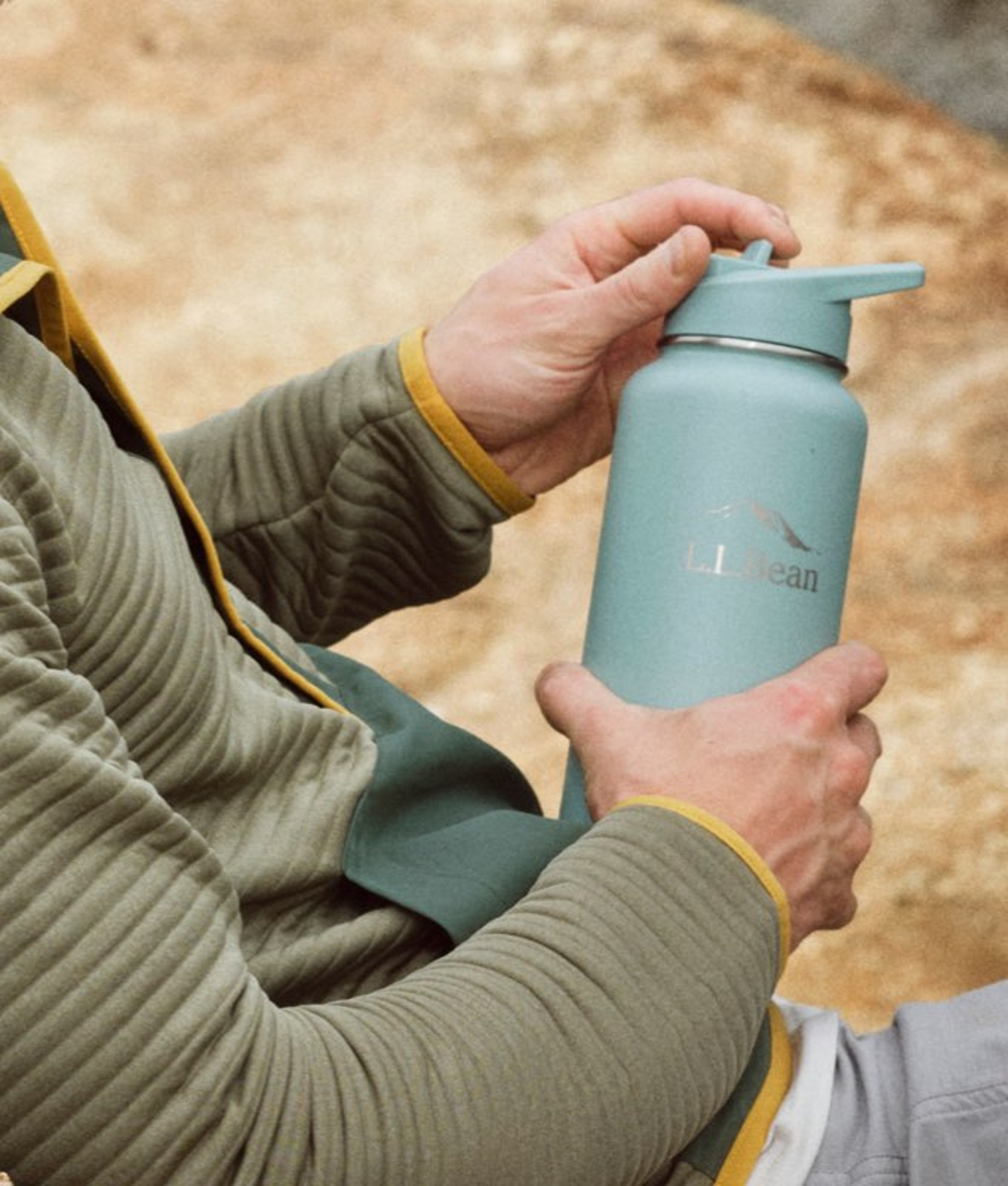 Person in green airlight top holding blue water bottle outdoors near sandy ground.
