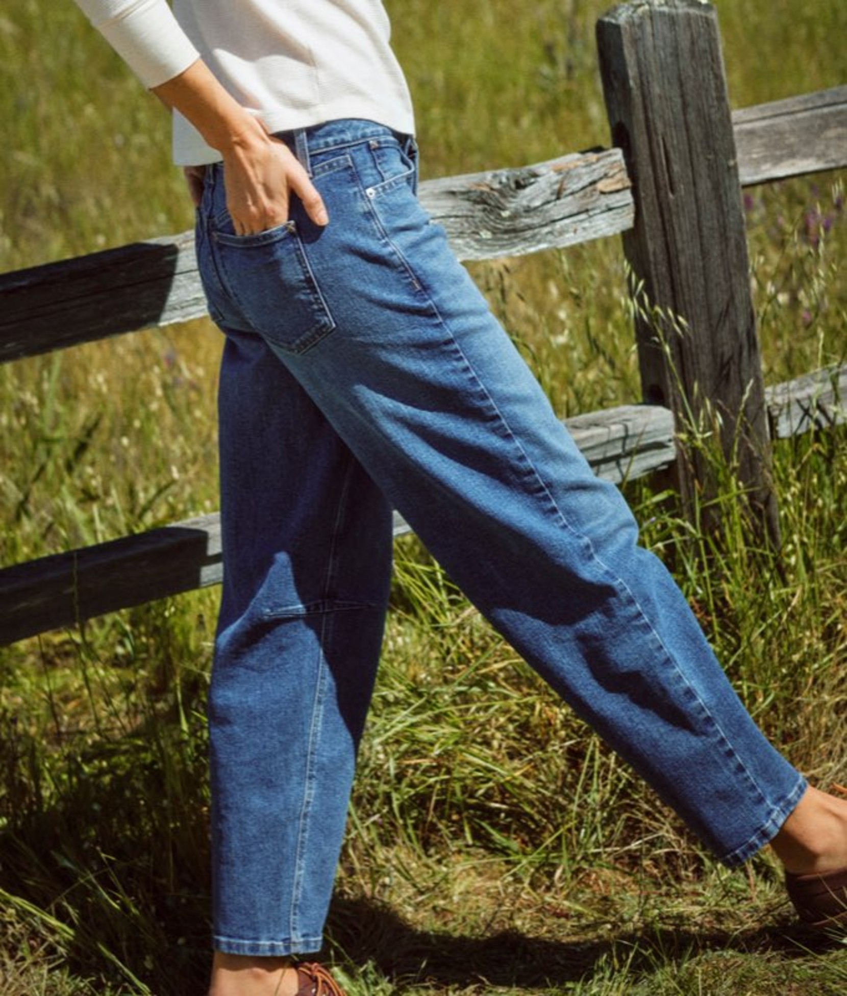 Woman wearing blue jeans walking along rustic wooden fence with tall grass behind it. 