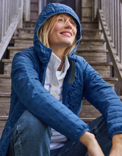 Person sits on wooden steps wearing a blue quilted hooded jacket and jeans.