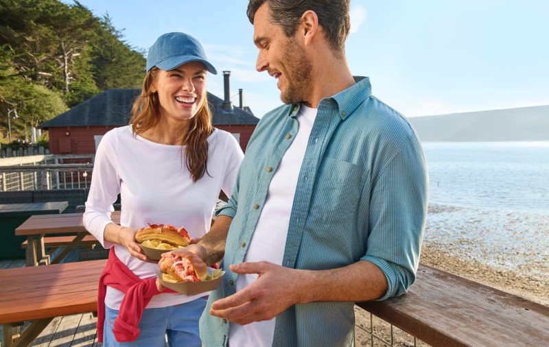 Two people holding lobster rolls on a wooden deck overlooking a beach and water, with picnic tables and buildings behind.