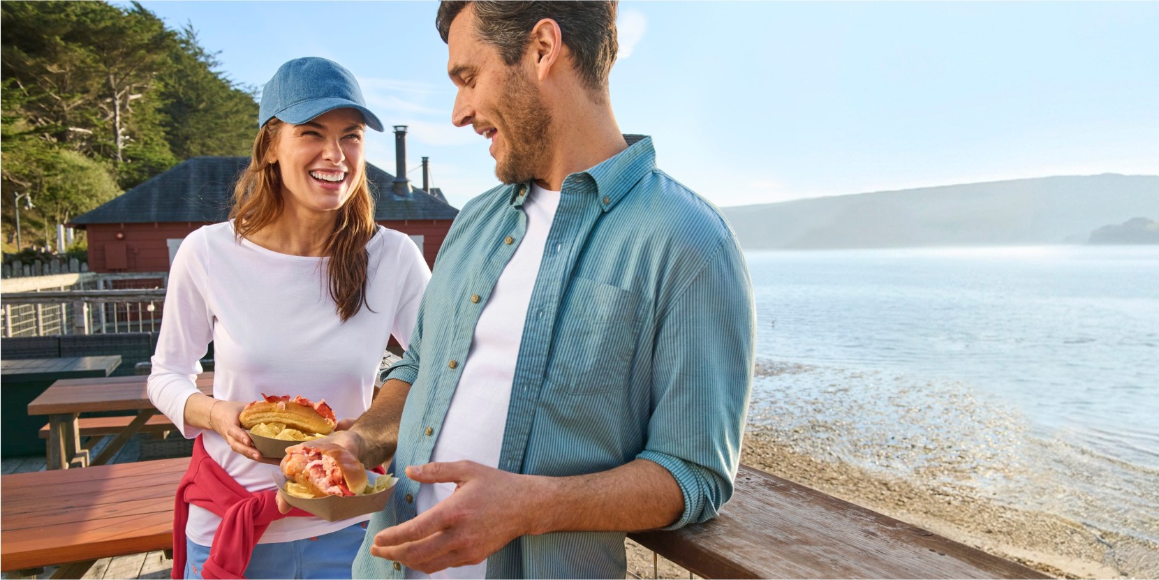 Two people holding lobster rolls on a wooden deck overlooking a beach and water, with picnic tables and buildings behind.