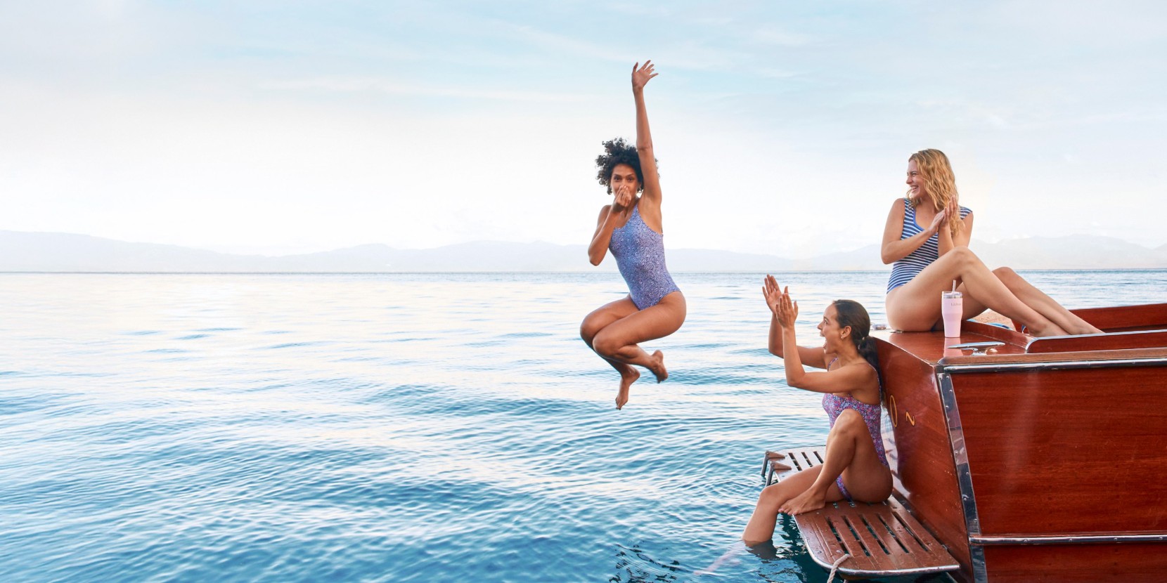 Three friends in swimsuits enjoy a boat day; one jumps into the blue water while two others cheer from a wooden deck.
