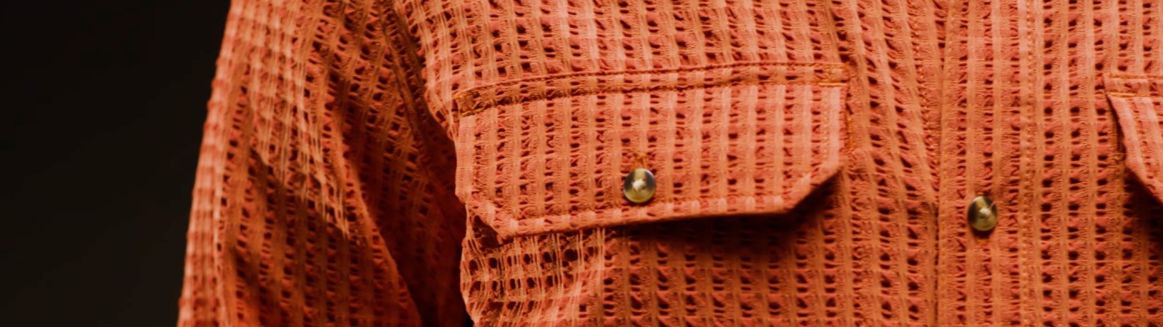 Close-up of a textured, rust-orange seersucker button-down shirt featuring chest pockets with small brown buttons.