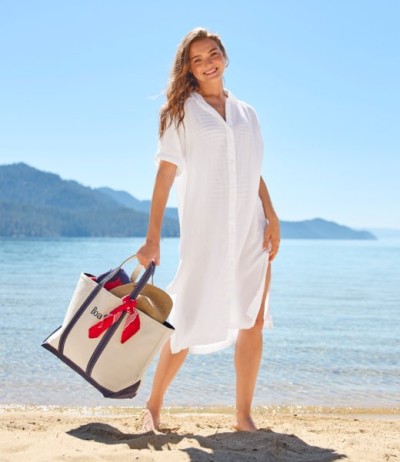 Person in white long-sleeve cover-up dress standing barefoot on a beach, holding a tote bag, water and hills behind.