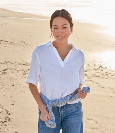Person in white gauze shirt and jeans standing on a sandy beach, holding a sweater, with shoreline and water in background.